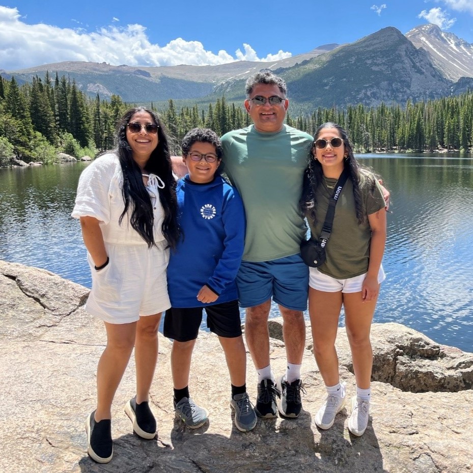 Ajit with his wife and children sanding in front of a lake with mountains and blue cloudy skies in the background.