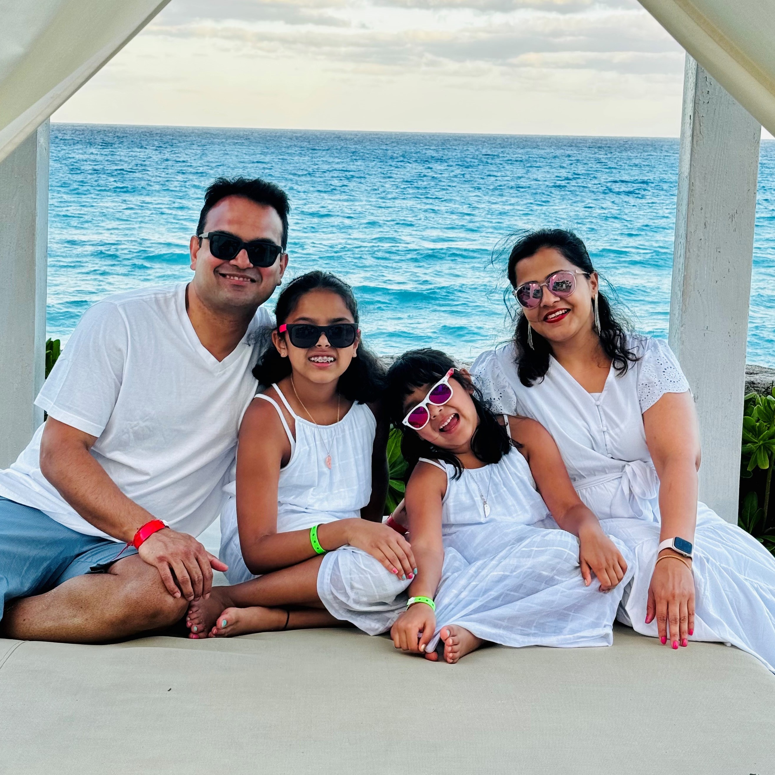 Neha with her husband and two daughters sitting on a large white cabana lounge chair on the beach with green foliage and the ocean in the background.