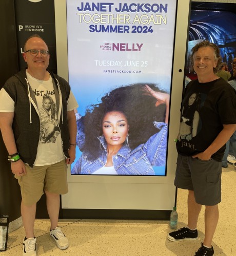 Ric Hunter stands next to a concert poster featuring singer Janet Jackson. Ric's husband stands opposite of Ric.