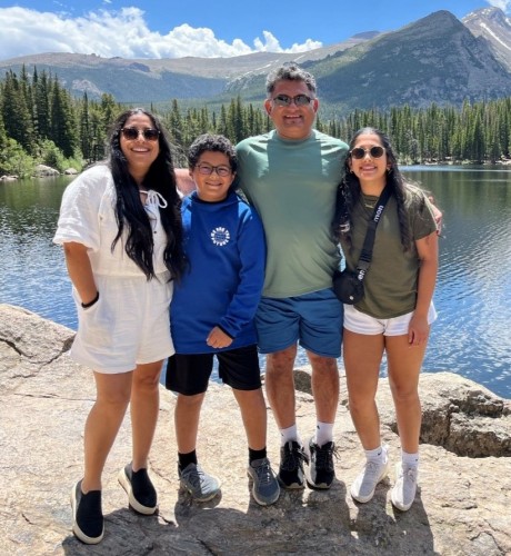 Ajit with his wife and children sanding in front of a lake with mountains and blue cloudy skies in the background.