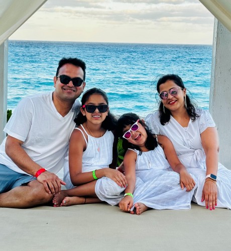 Neha with her husband and two daughters sitting on a large white cabana lounge chair on the beach with green foliage and the ocean in the background.