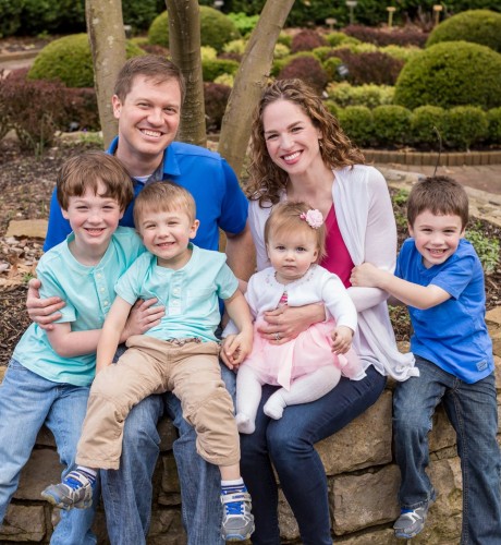 John, his wife, and their four children sit on a stone wall in a park and smile for a photo
