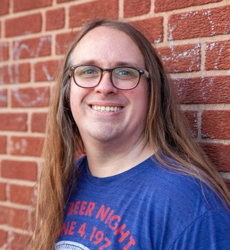 Jimmy smiles in a professional headshot taken against a red brick backdrop.