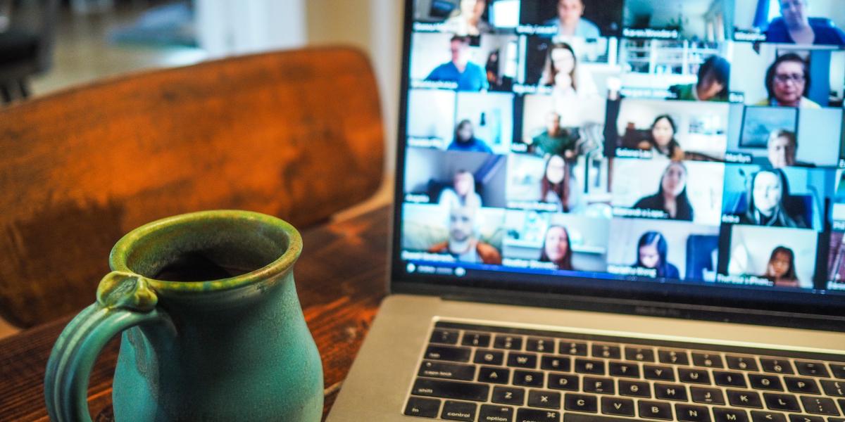 A mug sits next to a laptop, the screen showing a large virtual gathering on Zoom