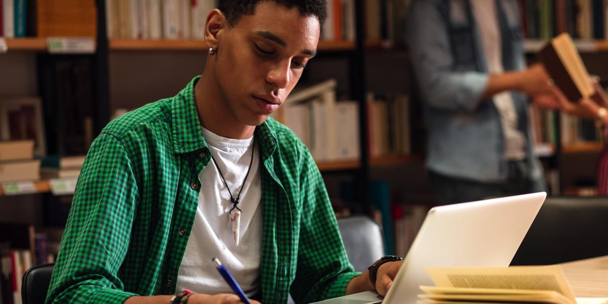 A student is in the library, working on an assignment using a laptop and a notebook.
