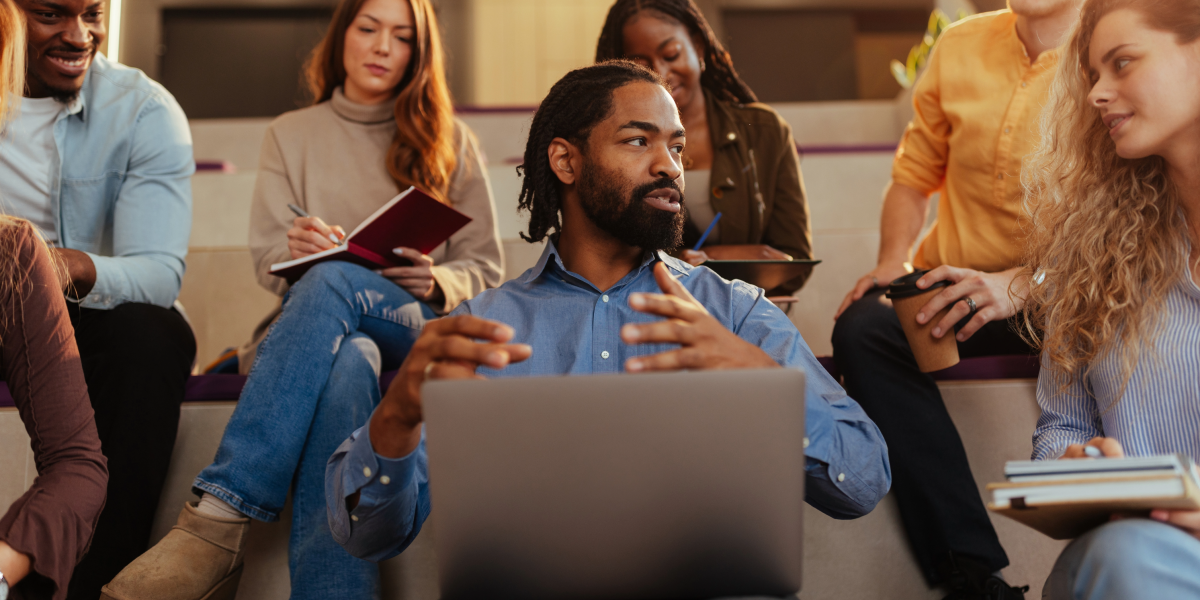 An instructor engages his students in discussion while working on a laptop