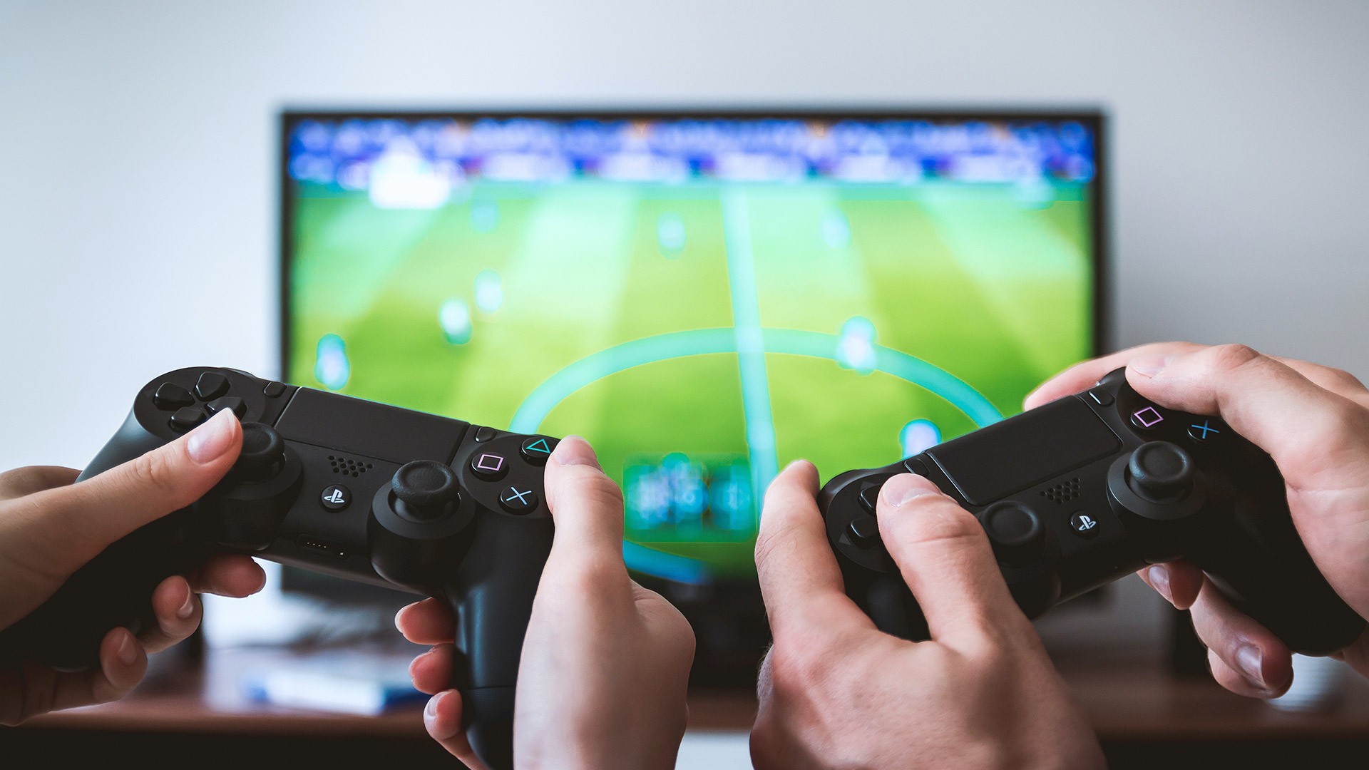 Hands holding controllers for a game console with a television in the background