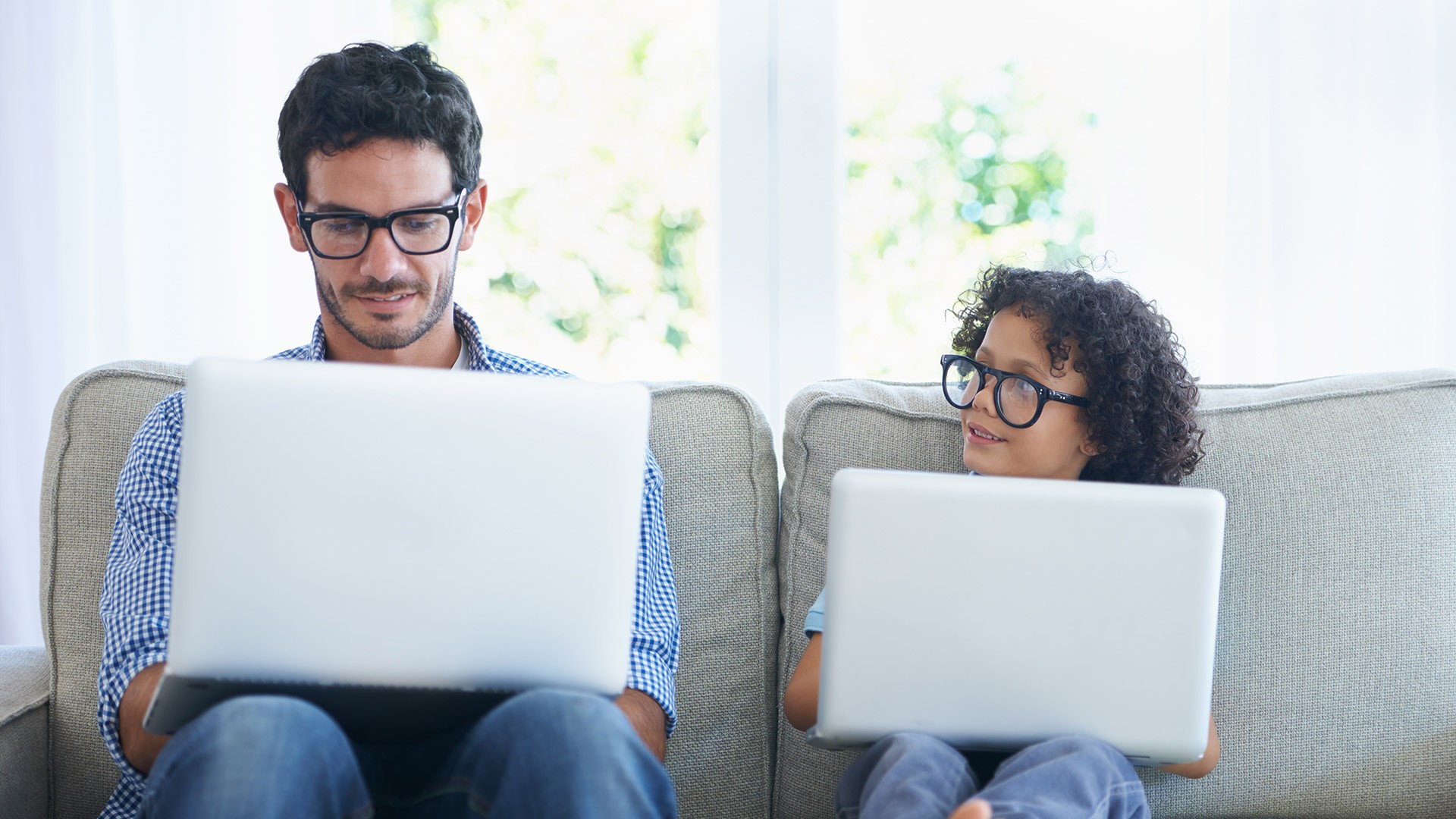 A man and a child sitting on a couch using laptops