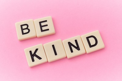 The phrase "Be Kind" spelled out on white tiles against a pink background.