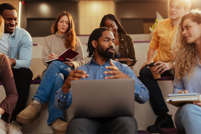 An instructor engages his students in discussion while working on a laptop