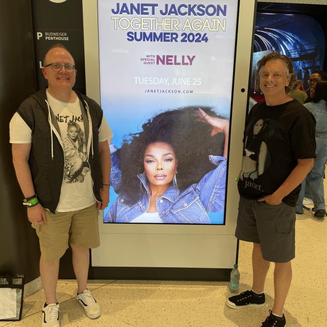 Ric Hunter stands next to a concert poster featuring singer Janet Jackson. Ric's husband stands opposite of Ric.