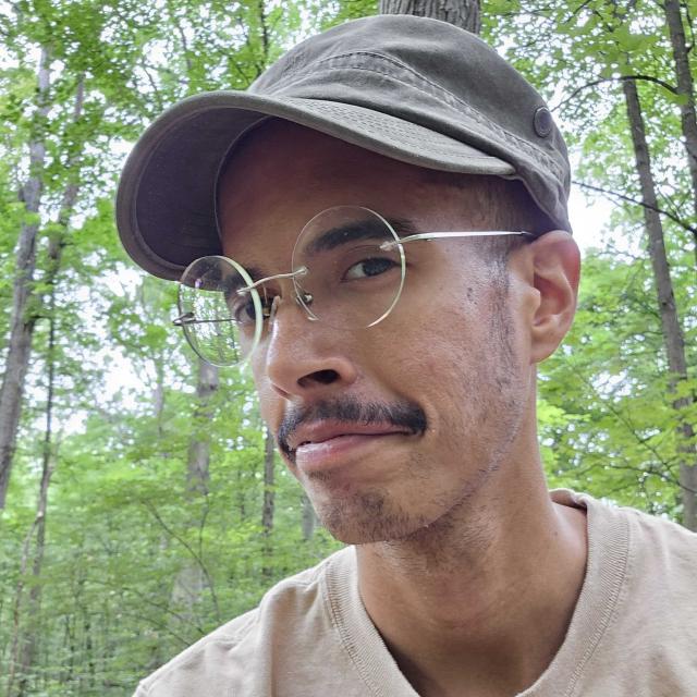 A man, Elliot Underwood, wears a baseball cap and rimless glasses and smirks at the camera. Elliot is outdoors in a woodlands setting.
