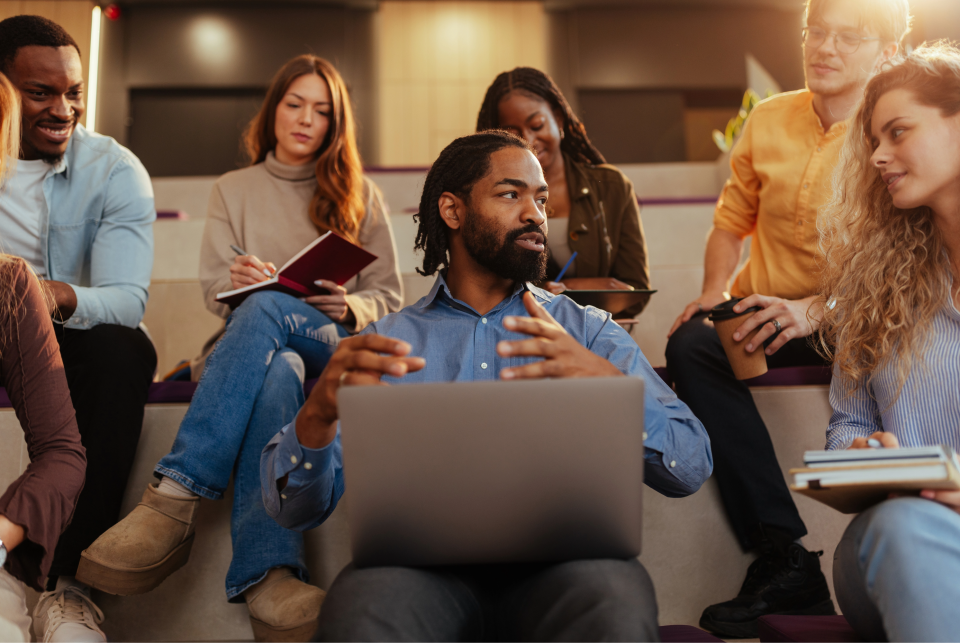 An instructor engages his students in discussion while working on a laptop